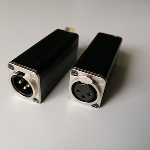 Puede incluir: Dos conectores XLR negros con extremos de metal plateado. Uno tiene tres pines, el otro tiene tres agujeros. Ambos tienen un cuerpo negro rectangular y están asegurados con tornillos pequeños. Estos conectores son para equipos de audio profesionales.