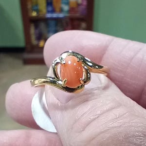 Puede incluir: Un anillo de oro con una piedra preciosa ovalada de color rosa engastada en un diseño de espiral.