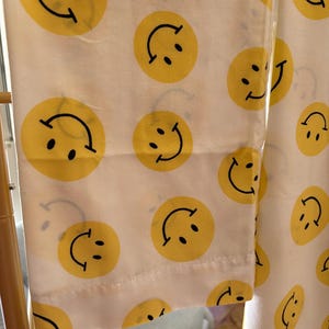 以下が含まれることがあります： 明るい黄色のスマイリーフェイスが繰り返しプリントされた白い布地。それぞれのスマイリーフェイスは、2つの黒い円形の目と口を表す湾曲した黒い線で構成されており、明るい色の表面全体に均等に配置されています。