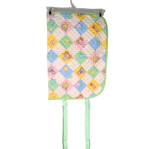 May include: A quilted fabric pad featuring a diamond pattern in pastel blue, pink, yellow, and green. The design includes small illustrations of children and balloons within the diamonds. The pad has a light green border and two long green ties.