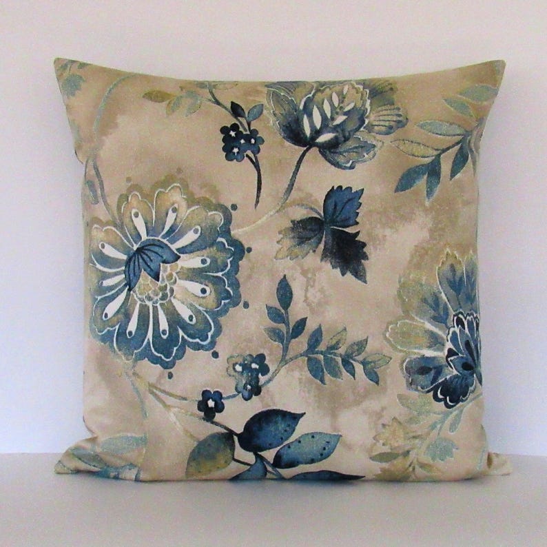 K&ouml;nnte beinhalten: Ein beigefarbenes Kissen mit einem blauen und gr&uuml;nen Blumendesign. Das Kissen ist quadratisch und aus einem weichen, bequemen Stoff gefertigt.