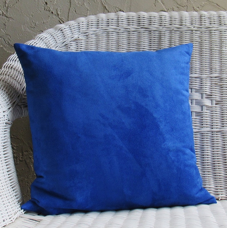 Op de afbeelding: Een koninklijk blauw su&egrave;de kussen met een zacht, gestructureerd oppervlak. Het kussen ligt op een witte rieten stoel.