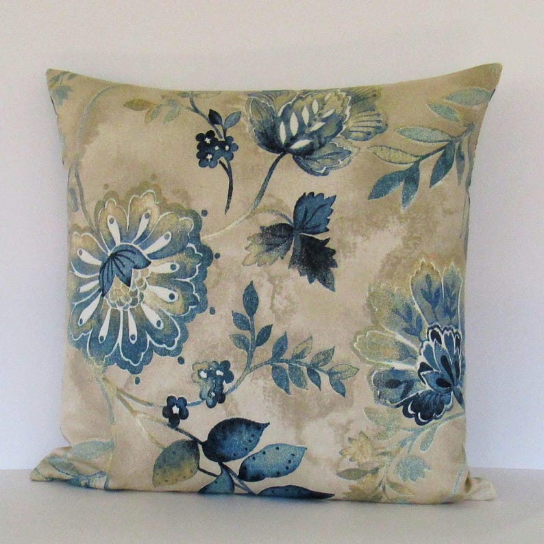 K&ouml;nnte beinhalten: Beigefarbener Kissenbezug mit einem blauen und gr&uuml;nen Blumenmuster. Das Kissen zeigt eine gro&szlig;e blaue Blume mit wei&szlig;en Akzenten in der Mitte.