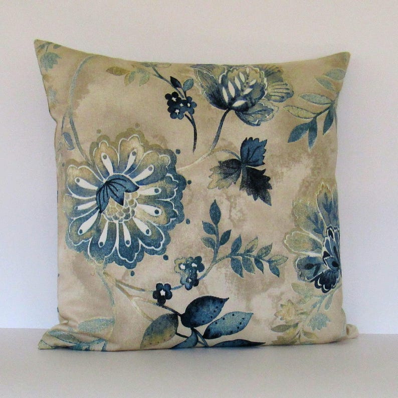 K&ouml;nnte beinhalten: Ein beiger Kissenbezug mit einem blauen und gr&uuml;nen Blumendesign. Das Kissen zeigt eine gro&szlig;e blaue Blume mit wei&szlig;en Akzenten und kleinere blaue Blumen im gesamten Design.