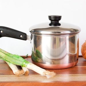May include: A stainless steel saucepan with a copper-colored base, a black handle featuring a metal hanging loop, and a matching stainless steel lid with a black knob. The pot is positioned next to green scallions and a whole yellow onion.