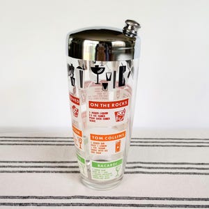 Puede incluir: Una coctelera vintage con tapa de acero inoxidable y cuerpo de vidrio transparente. El vidrio est&aacute; decorado con gr&aacute;ficos rojos y negros y recetas de c&oacute;cteles, incluyendo "Tom Collins" y "Bacardi". La coctelera tiene un colador incorporado.