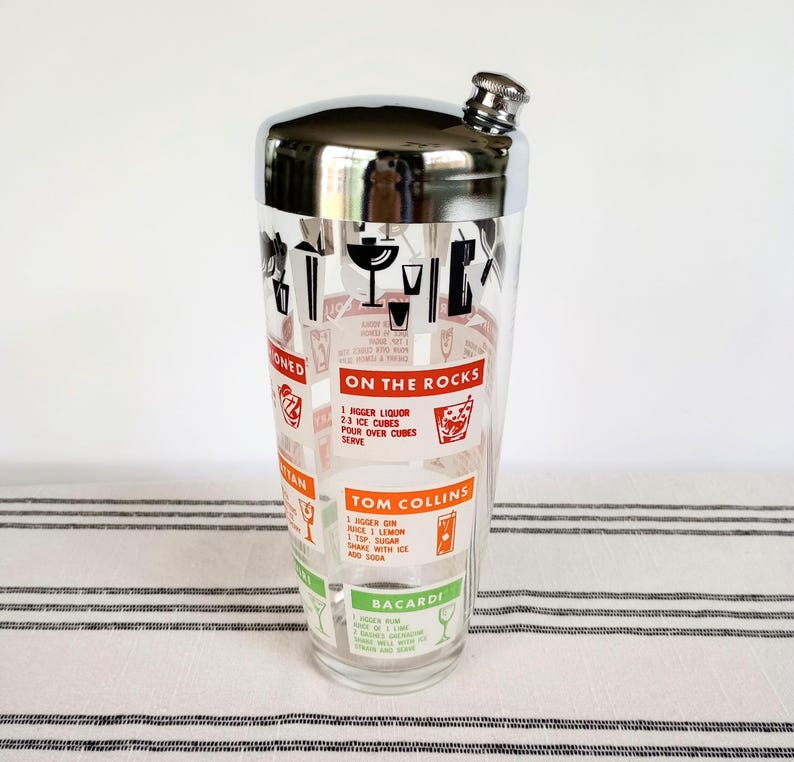 Puede incluir: Una coctelera vintage con tapa de acero inoxidable y cuerpo de vidrio transparente. El vidrio est&aacute; decorado con gr&aacute;ficos rojos y negros y recetas de c&oacute;cteles, incluyendo "Tom Collins" y "Bacardi". La coctelera tiene un colador incorporado.