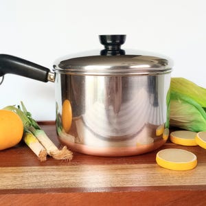 May include: A stainless steel pot with a black handle and lid, set on a wooden surface. Accompanied by a lemon, green onions, lettuce, and lemon slices. The pot is reflective, and the scene suggests a cooking theme.