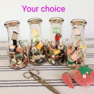 May include: Four clear glass milk-style bottles filled with assorted colorful sewing thread spools and various buttons. A pair of silver metal scissors and a red tomato-shaped pincushion with a small matching strawberry are placed in front.