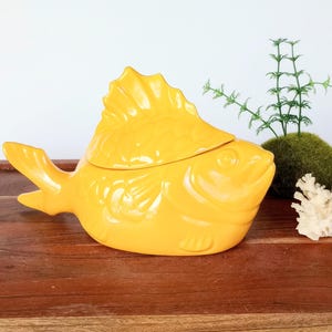 May include: A bright yellow ceramic fish-shaped container with a lid. The container has a detailed fin and tail design. Decorative elements include a small green plant and white coral on a wooden surface.