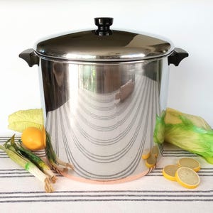 May include: A large, shiny stainless steel pot with a black handle and lid. The pot is surrounded by green vegetables, a yellow lemon, and green onions. The pot has a copper-colored bottom.