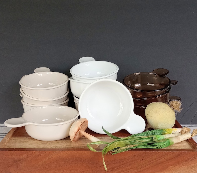 Pode incluir: Uma cole&ccedil;&atilde;o de tigelas de sopa de cer&acirc;mica creme, brancas e castanhas. Cada tigela tem uma al&ccedil;a. As tigelas est&atilde;o dispostas numa t&aacute;bua de madeira com vegetais decorativos, incluindo cebolinho e um cogumelo.