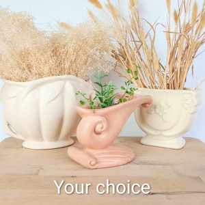 May include: Three ceramic planters in cream and peach tones. The left planter is round with vertical ridges, the center is a peach-colored scroll-shaped vessel with green foliage, and the right is a square cream planter with floral relief. Text: Your choice.