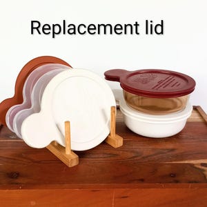 May include: A collection of round plastic replacement lids in white, translucent, and brown, featuring side tabs. The lids are organized on a small wooden rack, with one additional brown lid placed on a white bowl next to the rack. Text reads Replacement lid.