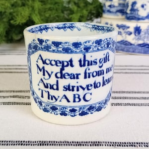 May include: A white ceramic cup with blue floral patterns and the text "Accept this gift My dear from me And strive to learn Thy ABC". The cup is part of a set of blue and white china.