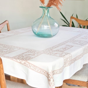 May include: A white rectangular tablecloth with a decorative border in a light brown color. A clear, light blue glass vase with dried plants sits in the center of the table. Wooden chairs surround the table.