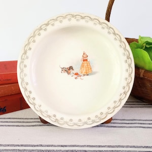 May include: A vintage white ceramic plate with a decorative border. The plate features a colorful illustration of a girl, a dog, and scattered objects. The plate is set on a wooden stand, with books and a basket in the background.