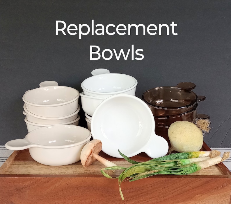 Pode incluir: Uma cole&ccedil;&atilde;o de tigelas de substitui&ccedil;&atilde;o nas cores branca, creme e castanha. As tigelas t&ecirc;m al&ccedil;as e est&atilde;o dispostas numa bandeja de madeira com vegetais decorativos. O texto na imagem diz "Replacement Bowls".