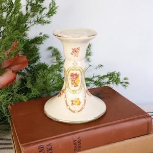 May include: A cream-colored ceramic candle holder featuring a flared base and top, decorated with delicate floral motifs in pink, yellow, and orange, accented by thin gold-toned trim along the edges and intricate decorative line work.