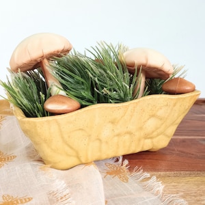 May include: A decorative arrangement featuring a yellow ceramic planter filled with faux greenery and several mushroom figurines. The mushrooms have tan caps and brown stems. The planter sits on a wooden surface.