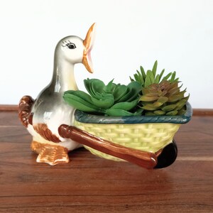 May include: A ceramic duck planter with a light green basket, holding faux succulents. The duck is gray, white, and brown, with an open beak. The basket has a dark blue trim and brown handles. A decorative piece for home decor.