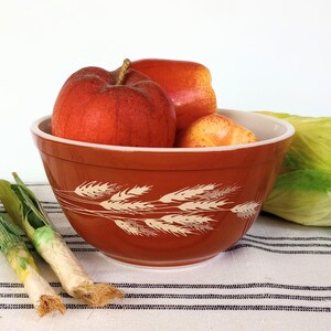 May include: A burnt orange mixing bowl featuring a white wheat stalk pattern around the exterior. The bowl is filled with two red apples and one yellow peach, with two green leeks resting on a white fabric surface with black stripes.