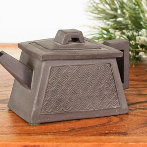 May include: A small, brown ceramic teapot with a square body and lid. The front of the teapot features a geometric pattern. The spout and handle are rectangular. The teapot is displayed on a wooden surface.