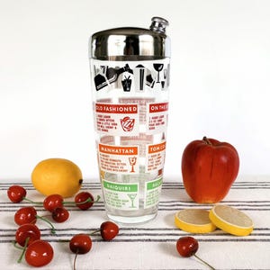 Puede incluir: Una coctelera vintage con tapa de acero inoxidable y cuerpo de vidrio con recetas de c&oacute;cteles. La coctelera est&aacute; decorada con ilustraciones y texto, incluyendo "Old Fashioned" y "Manhattan". Una manzana, rodajas de lim&oacute;n y cerezas est&aacute;n dispuestas cerca.