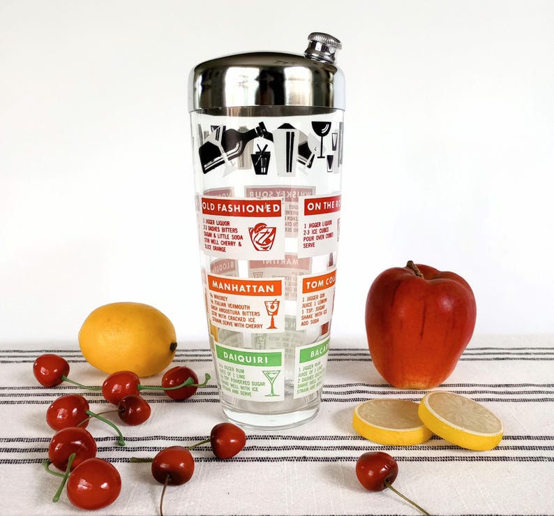 Puede incluir: Una coctelera vintage con tapa de acero inoxidable y cuerpo de vidrio con recetas de c&oacute;cteles. La coctelera est&aacute; decorada con ilustraciones y texto, incluyendo "Old Fashioned" y "Manhattan". Una manzana, rodajas de lim&oacute;n y cerezas est&aacute;n dispuestas cerca.