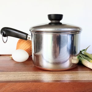 May include: A stainless steel saucepan with a black handle and lid. An egg, onion, and green onions are next to the pot on a wooden surface. The pot is reflective and appears to be new.