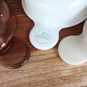 Pode incluir: Close-up de tr&ecirc;s tigelas Corning Ware Grab-It. Uma &eacute; castanha transparente, as outras s&atilde;o brancas e branco sujo. Cada tigela tem uma pega e as palavras "Grab It" e "Corning Ware" est&atilde;o impressas na parte inferior. As tigelas est&atilde;o numa superf&iacute;cie de madeira.