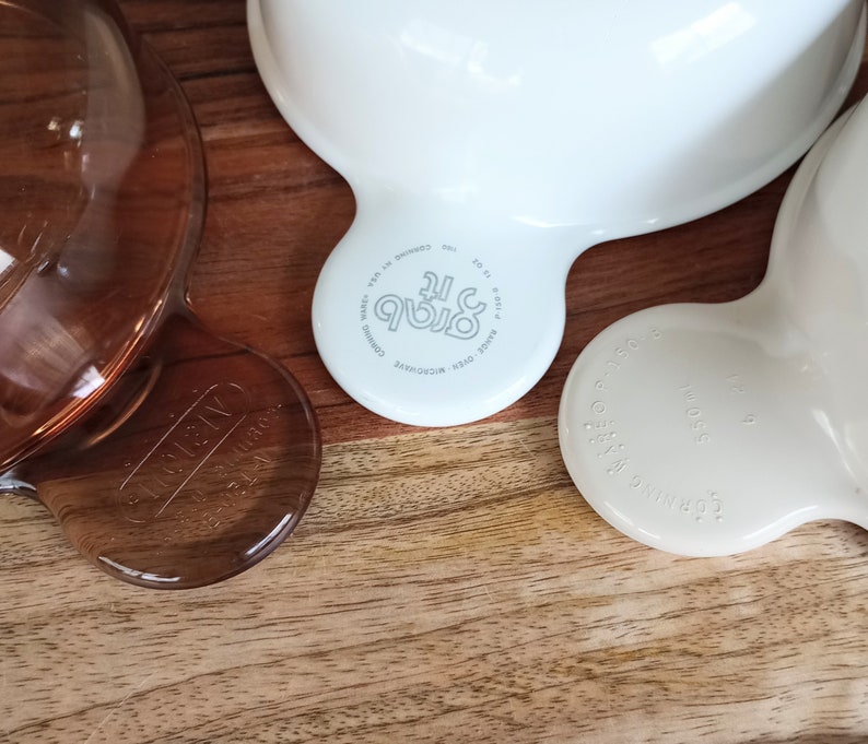 Pode incluir: Close-up de tr&ecirc;s tigelas Corning Ware Grab-It. Uma &eacute; castanha transparente, as outras s&atilde;o brancas e branco sujo. Cada tigela tem uma pega e as palavras "Grab It" e "Corning Ware" est&atilde;o impressas na parte inferior. As tigelas est&atilde;o numa superf&iacute;cie de madeira.