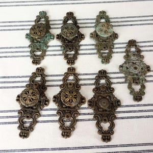 May include: A collection of seven ornate metal drawer pulls and backplates featuring a dark bronze finish with green patina accents. Each piece displays intricate scrollwork, a central circular knob or medallion, and mounting holes.