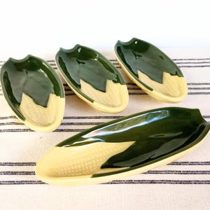 May include: A set of four ceramic dishes shaped like corn on the cob, featuring a glossy dark green glaze on the husk sections and a pale yellow finish on the textured kernel sections. The collection includes three smaller dishes and one larger dish.