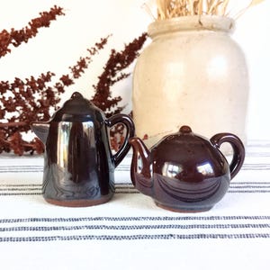 May include: Two small, dark brown ceramic teapots with handles and spouts. One is tall and slender, the other is round. They sit on a striped surface, with a large beige vase and dried plants in the background.