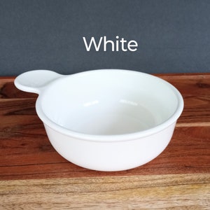 Pode incluir: Uma tigela de cer&acirc;mica branca com uma al&ccedil;a, contra um fundo escuro. A tigela &eacute; redonda com uma borda ligeiramente alargada e uma pequena al&ccedil;a em um lado. A palavra "White" &eacute; impressa acima da tigela.