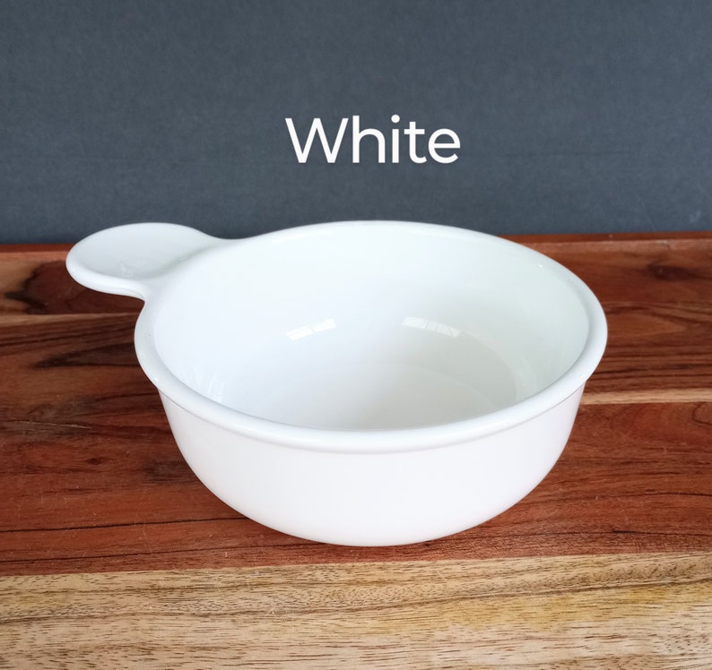 Pode incluir: Uma tigela de cer&acirc;mica branca com uma al&ccedil;a, contra um fundo escuro. A tigela &eacute; redonda com uma borda ligeiramente alargada e uma pequena al&ccedil;a em um lado. A palavra "White" &eacute; impressa acima da tigela.