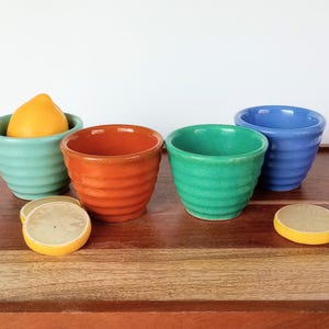 May include: A collection of colorful ceramic bowls in shades of teal, orange, green, and blue, with a lemon and lemon slices. The bowls have a ribbed design and are arranged on a wooden tray.
