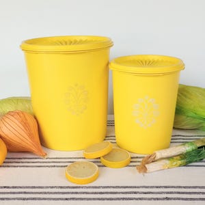 May include: Two bright yellow vintage food storage containers with lids. Each container has a white floral design. The containers are on a striped cloth with lemons, onions, and cabbage.
