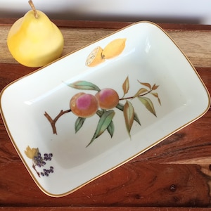 May include: A rectangular white ceramic dish with a gold rim, featuring a hand-painted design of peaches, lemons, and grapes. A yellow pear sits to the left of the dish. The fruit illustrations are detailed with green leaves and brown branches.