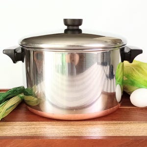 May include: A stainless steel stockpot with a polished finish, a matching lid, and black heat-resistant side handles. The base of the pot features a visible copper-colored band. The pot is cylindrical with a flat lid and a central black knob.