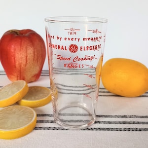 May include: A clear General Electric measuring glass with red text and measurement markings. The glass is next to a red apple, a yellow lemon, and three lemon slices. The text on the glass reads "Best by every measure" and "Speed Cooking Ranges."