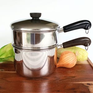 May include: A stainless steel double boiler pot with a matching lid. The top section features a black handle, while the bottom section has a dark brown handle. Both handles include a small metal loop at the end for hanging storage purposes.
