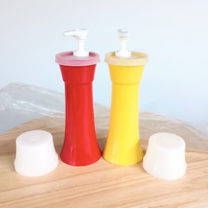 May include: Two plastic condiment dispensers, one red and one yellow, with white pump tops and matching lids. The dispensers are on a wooden surface. Two white lids are also present.