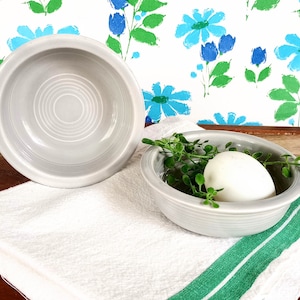 May include: Two gray ceramic bowls, one with a white egg and green sprigs inside. The bowls have a ridged design and are on a white towel with green stripes.