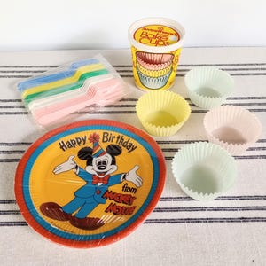 May include: A collection of party supplies, including a Mickey Mouse birthday plate, colorful forks, cupcake liners, and a container of baking cups. The plate features Mickey Mouse with the text "Happy Birthday from Mickey Mouse."