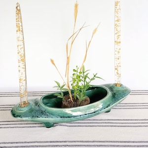 May include: A teal ceramic centerpiece with a boat-like shape, holding greenery and dried grasses. Two clear, gold-flecked glass candle holders flank the centerpiece. The arrangement sits on a striped white and blue surface.