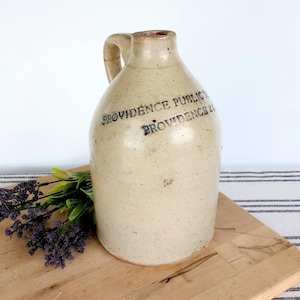 May include: A beige stoneware jug with a handle, featuring the text "PROVIDENCE PUBLIC" and "PROVIDENCE 21" in dark blue. The jug is on a wooden cutting board with purple and green floral accents.