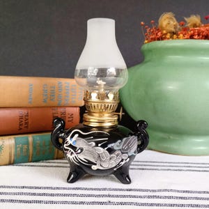 May include: A small oil lamp with a black ceramic base featuring a white and grey dragon design, three legs, and two side handles. The lamp has a gold-toned metal burner assembly and a clear glass chimney with a frosted white top section.