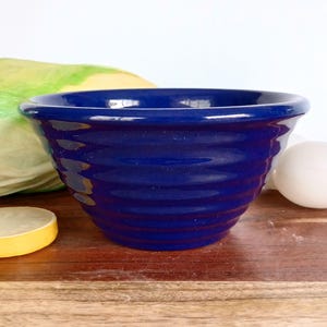 May include: A deep blue ceramic bowl with a ribbed exterior. The bowl is set on a wooden surface, with a yellow object and a head of lettuce visible in the background. The bowl is empty.